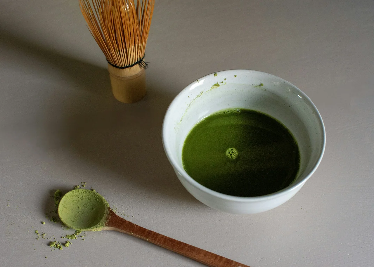 A wooden spoon of matcha powder beside a bowl of whisked matcha and a bamboo whisk