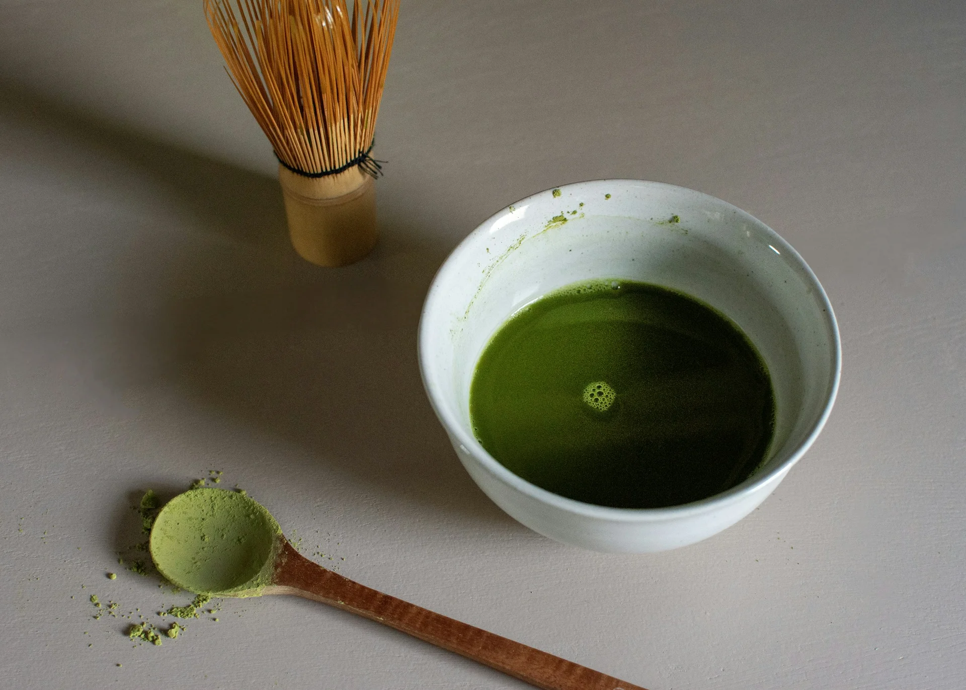 A wooden spoon of matcha powder beside a bowl of whisked matcha and a bamboo whisk